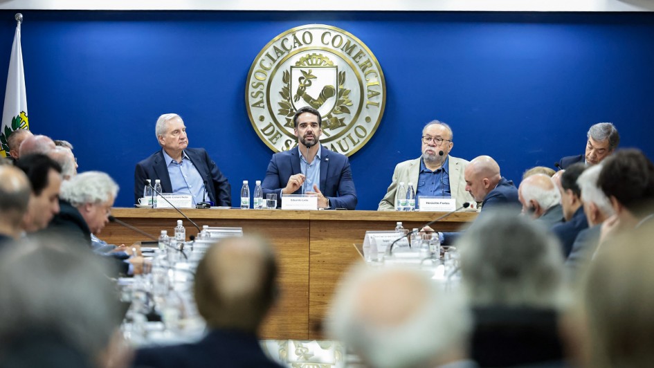 Foto de Leite sentado numa bancada ao lado de dois homens. Ao fundo, há uma parede azul com o símbolo, de cor branca e redondo, da Associação Comercial de SP. Leite fala ao microfone. No primeiro plano da foto, é possível ver as cabeças das pessoas que assistem. 