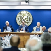 Foto de Leite sentado numa bancada ao lado de dois homens. Ao fundo, há uma parede azul com o símbolo, de cor branca e redondo, da Associação Comercial de SP. Leite fala ao microfone. No primeiro plano da foto, é possível ver as cabeças das pessoas que assistem. 