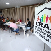 Foto mostra grupo de oito mulheres sentadas ao redor de uma grande mesa retangular e praticando crochê