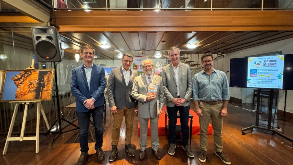 A imagem mostra cinco homens em pé, posando para a foto. No lado esquerdo da imagem, há uma fotografia em um cavalete. No lado direito, há um painel com uma apresentação sobre os 400 anos das Missões. 