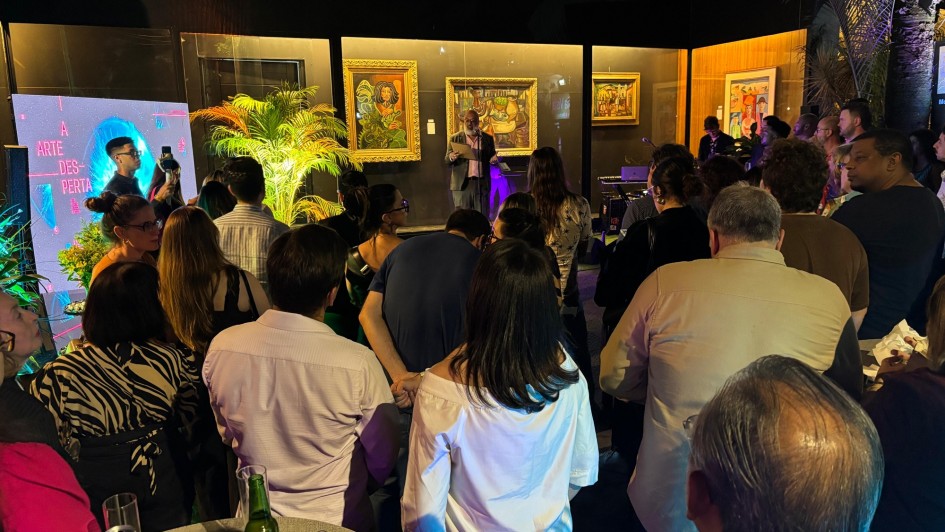 Foto mostra o lançamento da programação da Noite dos Museus, com público reunido em frente a um palco interno decorado com plantas e obras de arte expostas nas paredes. A iluminação em tons quentes e o painel com o slogan “A arte desperta” compõem o cenário, enquanto um orador faz pronunciamento diante da plateia atenta.
