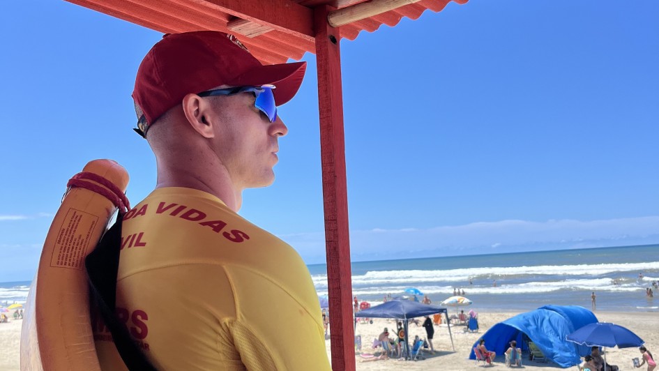 A imagem mostra um homem com uniforme de guarda-vidas civil em cor amarela e vermelha. Ele está de perfil, de boné e de óculos escuros olhando, em direção ao mar ao fundo da imagem. 