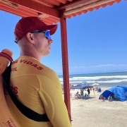 A imagem mostra um homem com uniforme de guarda-vidas civil em cor amarela e vermelha. Ele está de perfil, de boné e de óculos escuros olhando, em direção ao mar ao fundo da imagem. 