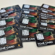 Uma foto em ângulo inclinado mostrando um grupo de cartões de plástico de forma retangular (semelhantes a cartões de crédito), sobrepostos em uma superfície clara, formando vários montes. Os cartões são predominantemente pretos e apresentam um design gráfico vibrante com gradientes de cores (vermelho, amarelo e verde) e a imagem de uma estátua ou monumento.

No canto inferior, em letras grandes e brancas, está escrito "TCHÊ CARD" e logo abaixo "AGÊNCIA DE TURISMO". Cada cartão também possui um QR Code grande e preto, com a inscrição "VIVA O RS" logo abaixo. A parte superior do cartão exibe, de forma discreta, a inscrição "TCHÊ CARD". Os cartões são o "Tchê Card", um produto de turismo do Rio Grande do Sul.