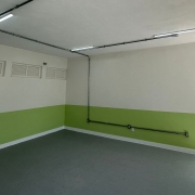 A imagem mostra uma sala de aula vazia, com as paredes nas cores branca e verde. O piso é cinza e há lâmpadas iluminando o ambiente. 