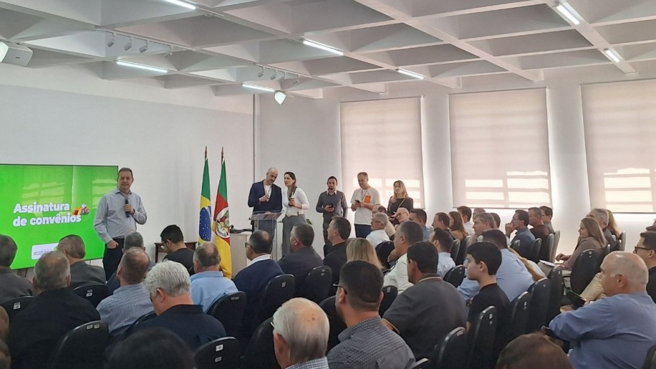 Autoridades estaduais e municipais participam de uma cerimônia de assinatura de convênios em um auditório. A imagem mostra o evento com plateia cheia e telão exibindo a mensagem “Assinatura de convênios”, ao lado das bandeiras do Brasil e do Rio Grande do Sul. Representantes do governo discursam à frente, em um ambiente iluminado e formal.