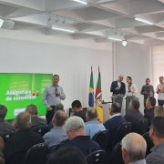 Autoridades estaduais e municipais participam de uma cerimônia de assinatura de convênios em um auditório. A imagem mostra o evento com plateia cheia e telão exibindo a mensagem “Assinatura de convênios”, ao lado das bandeiras do Brasil e do Rio Grande do Sul. Representantes do governo discursam à frente, em um ambiente iluminado e formal.