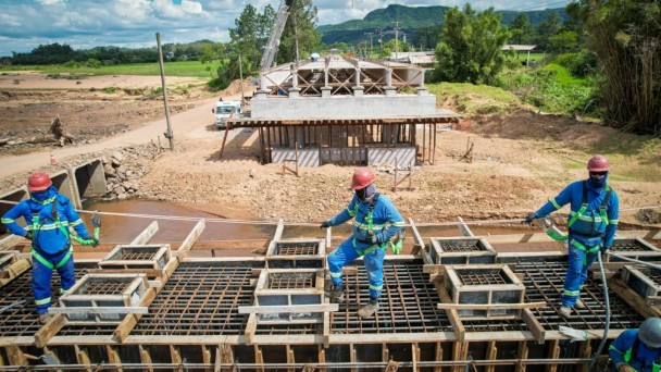 Três trabalhadores com uniformes azuis e capacetes vermelhos estão sobre uma estrutura de concreto e aço em construção de uma ponte. Eles usam cintos de segurança e estão posicionados sobre vigas e armações metálicas. Ao fundo, há outra parte da ponte em fase mais avançada, com colunas e lajes já concretadas. 