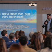 Uma foto interna em close-up de uma palestra ou evento em um auditório ou sala de conferências. Um homem (Gabriel Souza), no lado direito, está de pé no palco, vestindo um blazer escuro sobre uma camisa social azul-clara, e falando em um microfone que segura com a mão direita.

Atrás e à esquerda dele, há uma grande tela de projeção. O slide exibido na tela é dominado pela imagem de uma paisagem urbana à beira-mar (possivelmente Porto Alegre). No topo do slide, o texto em letras grandes e coloridas diz "RIO GRANDE DO SUL DO FUTURO" e, abaixo, "TRANSFORMAÇÃO, RECONSTRUÇÃO E DESENVOLVIMENTO".

Em primeiro plano, a parte superior da cabeça e dos ombros de vários membros da audiência, que estão de costas para a câmera, preenche o terço inferior da imagem. No canto superior direito, há uma placa com o nome "AMCHAM". A iluminação é focada no palestrante e no slide.