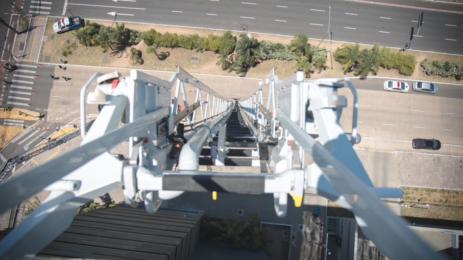 A imagem apresenta uma perspectiva vertical a partir do topo de uma autoescada mecânica, mostrando a estrutura metálica da escada em primeiro plano. Abaixo, é possível ver uma avenida larga com faixas de trânsito e algumas árvores.