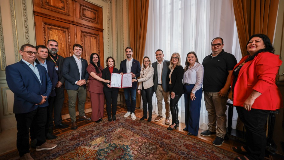 Uma foto colorida, tirada em ambiente interno que parece ser um gabinete ou sala de reuniões formal, possivelmente em um palácio de governo, devido à arquitetura e decoração. Um grupo de doze pessoas, composto por homens e mulheres, está posicionado no centro da sala, em pé sobre um tapete estampado.

No centro do grupo, um homem (aparentemente o governador) e uma mulher estão segurando um documento (possivelmente um ato de autorização ou convênio), indicando uma assinatura ou entrega formal.

Pessoas: Estão vestidas com trajes sociais ou casuais de negócios. O homem no centro-direita (governador) veste blazer e camisa polo. Outros homens usam ternos, blazers ou camisas sociais. As mulheres usam blazers, calças sociais e blusas.

Cenário: A sala tem um alto pé-direito. Ao fundo, uma grande porta de madeira escura com painéis esculpidos ocupa a parede central. À direita, janelas altas estão cobertas por cortinas longas e pesadas, de cor bege ou dourada, que permitem a entrada de luz natural. O piso é coberto por um grande tapete oriental, ricamente estampado em tons de bordô, azul-marinho e bege. A iluminação é clara e uniforme.

A pose é formal, com todos olhando para a câmera e sorrindo, sugerindo um momento de celebração ou registro de um acordo importante.