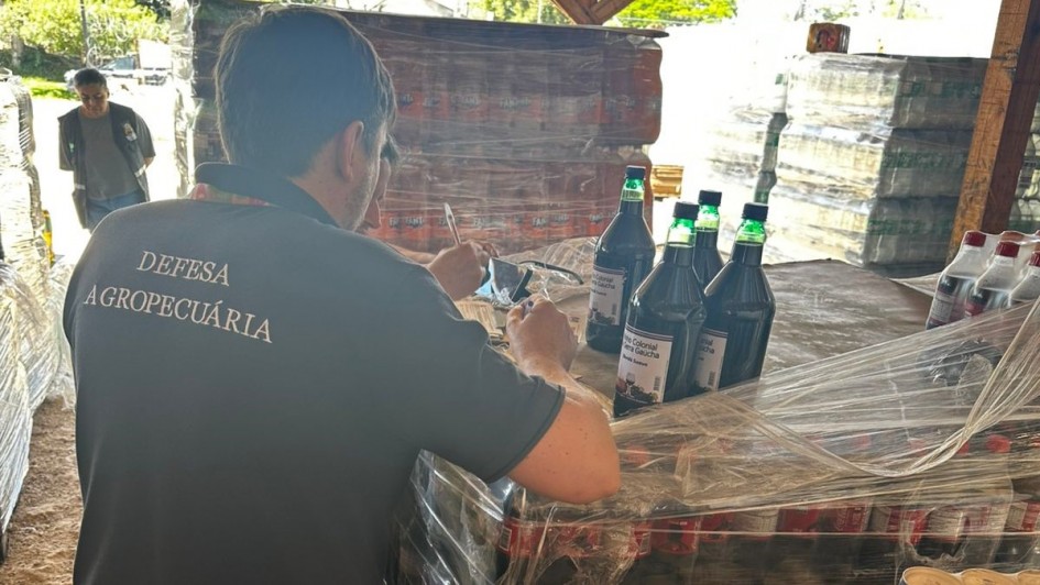 A imagem mostra um homem de costas, com camiseta preta em que se lê "Defesa Agropecuária". Ao lado dele, há fardos de bebidas e, em destaque, quatro litros de vinho colonial. 
