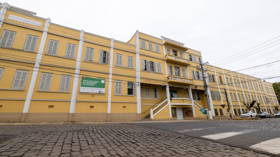 A imagem mostra a fachada do prédio histórico conhecido como Castelinho, em tom amarelo, localizado em rua de paralelepípedos. Na lateral do edifício há uma placa verde com os dizeres “Mais uma obra do Governo do Estado”. O prédio tem janelas com venezianas brancas e uma escadaria central, representando o investimento em restauração e preservação de patrimônios no Vale do Taquari.