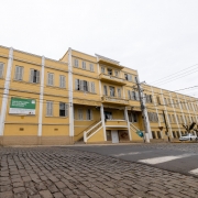 A imagem mostra a fachada do prédio histórico conhecido como Castelinho, em tom amarelo, localizado em rua de paralelepípedos. Na lateral do edifício há uma placa verde com os dizeres “Mais uma obra do Governo do Estado”. O prédio tem janelas com venezianas brancas e uma escadaria central, representando o investimento em restauração e preservação de patrimônios no Vale do Taquari.