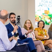 A imagem mostra o governador Eduardo Leite falando ao microfone. Ao lado dele, há outras quatro pessoas sentadas, compondo uma mesa-redonda. Ao fundo, há um painel com uma apresentação. 