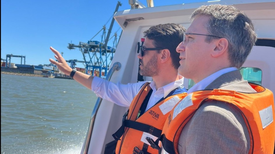 Foto de dois homens a céu aberto, navegando num barco. Eles aparecem de lado, trajando coletes laranjas de boiar. Um deles faz um gesto com o braço direito erguido na direção de um lugar fora do quadro, para o qual os dois olham.