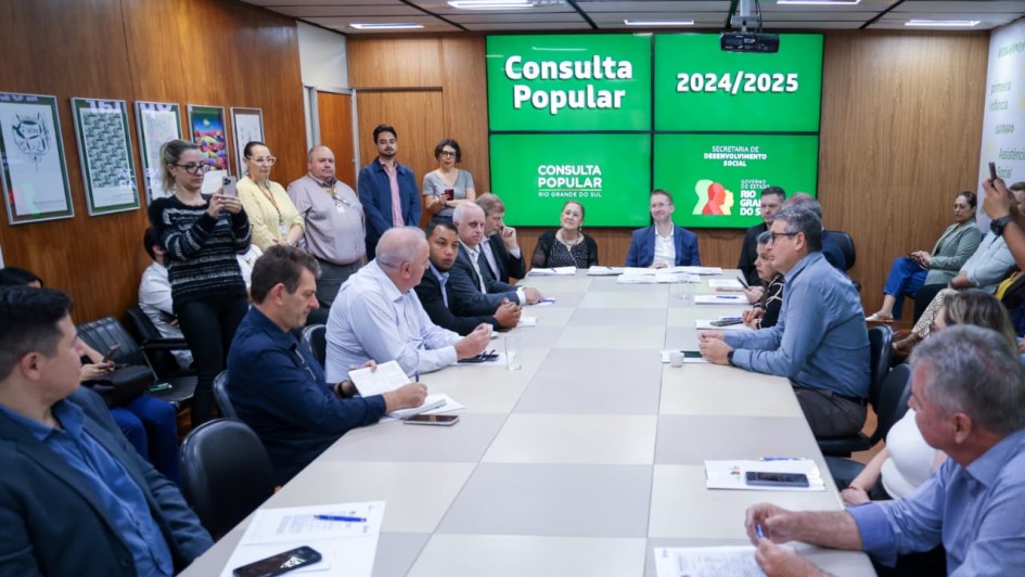 A imagem mostra diversas pessoas sentadas em uma mesa de reuni&otilde;es retangular. Na ponta da mesa, est&aacute; o secret&aacute;rio de Desenvolvimento Social. Ao fundo, em um tel&atilde;o de fundo verde, l&ecirc;-se "Consulta Popular 2024/2025" e a marca do governo do Estado do Rio Grande do Sul.