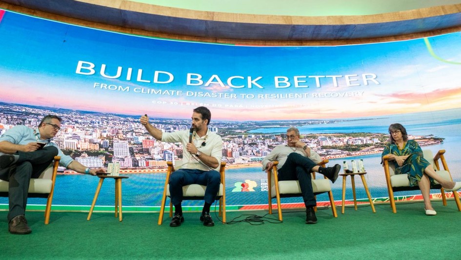 Painel com quatro participantes sentados em poltronas, diante de um grande telão com o título “BUILD BACK BETTER – From Climate Disaster to Resilient Recovery”. O ambiente tem iluminação verde e visual moderno. O governador Eduardo Leite aparece ao centro, segurando um microfone e gesticulando enquanto fala ao público. 