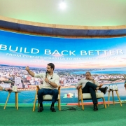 Painel com quatro participantes sentados em poltronas, diante de um grande telão com o título “BUILD BACK BETTER – From Climate Disaster to Resilient Recovery”. O ambiente tem iluminação verde e visual moderno. O governador Eduardo Leite aparece ao centro, segurando um microfone e gesticulando enquanto fala ao público. 
