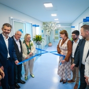 Foto de sete pessoas dentro do hospital descerrando a faixa de inauguração, de cor azul. Elas se encontram em um ambiente claro, de piso azul, e sorriem