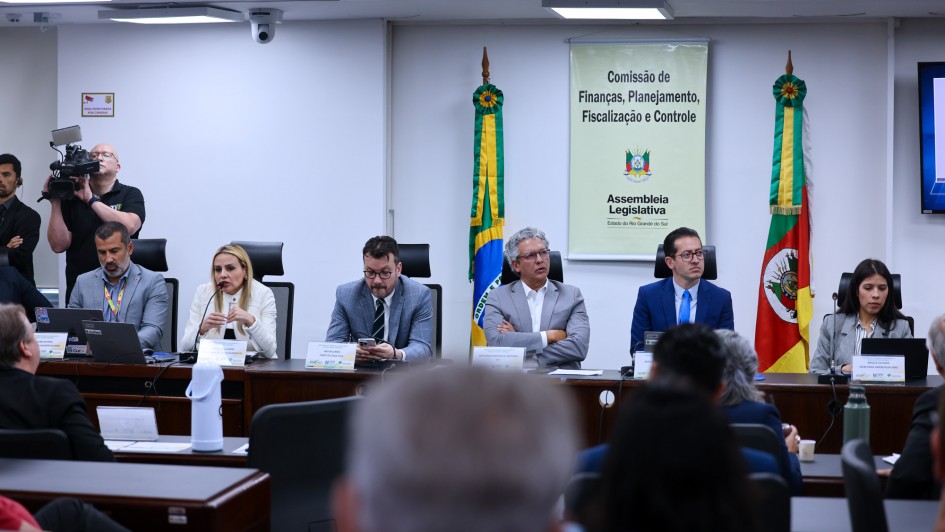 A imagem mostra uma reunião de comissão em um ambiente interno.

Pano de Fundo: No centro, há um banner vertical pendurado com o título "Comissão de Finanças, Planejamento, Fiscalização e Controle" seguido pelo brasão do estado e o texto "Assembleia Legislativa - Estado do Rio Grande do Sul". O banner está ladeado por duas bandeiras, possivelmente do Brasil e do Rio Grande do Sul.

Mesa Principal: Seis pessoas estão sentadas lado a lado em uma mesa, voltadas para o público (que está desfocado em primeiro plano).

Três homens à esquerda: O primeiro (esquerda) veste um terno cinza-claro. O segundo (centro) veste um blazer cinza-escuro e gravata. O terceiro (direita) veste um terno cinza-claro e está ligeiramente de lado.

Três pessoas à direita: O quarto (centro-direita) veste um terno azul-marinho. A quinta (direita) é uma mulher vestindo blazer branco, e a última pessoa visível na mesa (extrema direita) é uma mulher de blazer cinza.

Primeiro Plano: A visão da câmera está atrás de alguns membros da audiência (cabeças e costas desfocadas) que estão sentados em mesas, voltados para a mesa principal.

Lateral Esquerda: Um cinegrafista (homem de camiseta preta) está em pé à esquerda, operando uma câmera de vídeo profissional em um tripé.

A foto parece registrar uma sessão da Comissão de Finanças, Planejamento, Fiscalização e Controle da Assembleia Legislativa do Rio Grande do Sul, com membros da comissão sentados à mesa principal e participantes na audiência.