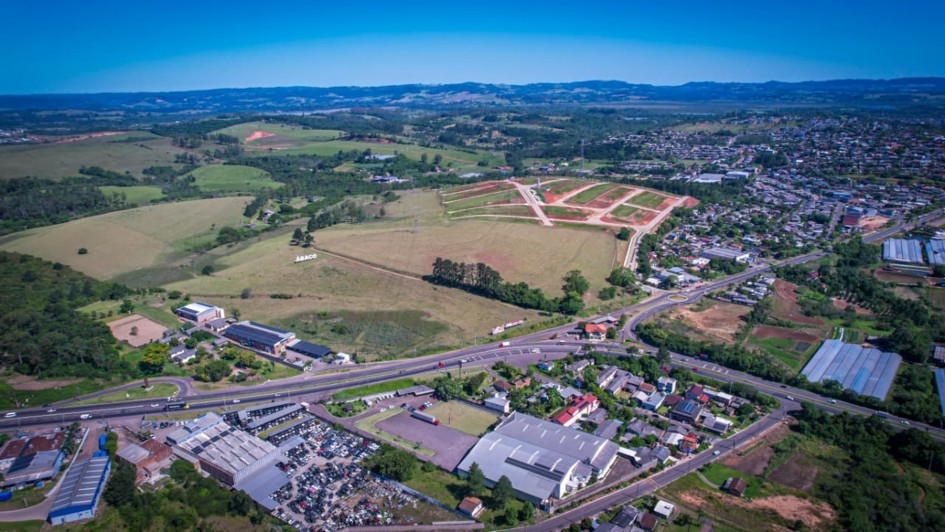 A imagem mostra uma vista aérea de uma área semiurbana. Em primeiro plano, há uma rodovia com veículos e prédios industriais e comerciais ao redor. Mais ao fundo, aparecem grandes áreas verdes e campos, com algumas construções espalhadas. À direita, há um bairro com muitas casas e ruas organizadas. No horizonte, vê-se colinas e vegetação sob um céu azul claro.