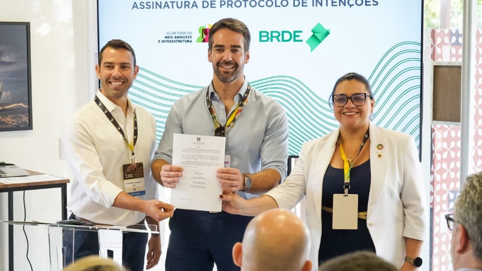 A imagem mostra três pessoas sorrindo, posando em pé para a foto. No meio, está o governador Eduardo Leite, segurando e exibindo um documento assinado. 