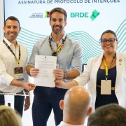 A imagem mostra três pessoas sorrindo, posando em pé para a foto. No meio, está o governador Eduardo Leite, segurando e exibindo um documento assinado. 