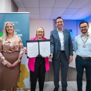 A imagem mostra quatro pessoas adultas em pé, lado a lado, em um ambiente interno, posando para uma foto oficial após a assinatura de um documento.

Pessoas (da esquerda para a direita):

Mulher: Veste uma blusa estampada em tons de pêssego e marrom sobre uma saia marrom-clara. Ela é loira, com um sorriso voltado para a câmera.

Mulher (Central): Veste uma jaqueta de cor rosa choque sobre calças pretas. Ela usa óculos e segura um documento dobrado (possivelmente um contrato ou convênio) em uma pasta preta, apresentando o papel aberto para a câmera.

Homem: Veste um terno cinza escuro sobre uma camisa branca sem gravata. Ele é moreno, com cabelo curto, sorrindo para a câmera.

Homem (Extrema Direita): Veste uma camisa social azul-clara e calças jeans escuras. Ele usa óculos e um crachá pendurado em um cordão laranja/vermelho.

Fundo:

No canto esquerdo, há um banner vertical turquesa (ou azul esverdeado) com texto parcial visível que inclui as palavras "SECRETARIA DA", "GOVERNO ESTADO" e "GRANDE".

No centro e à direita, a parede de fundo é clara, com um segundo banner ou painel no canto superior direito que mostra parte de um logotipo azul e branco, possivelmente institucional ("TAI S I").

No canto inferior esquerdo, parte de um kit de bateria (tambores vermelhos e brancos) é visível.