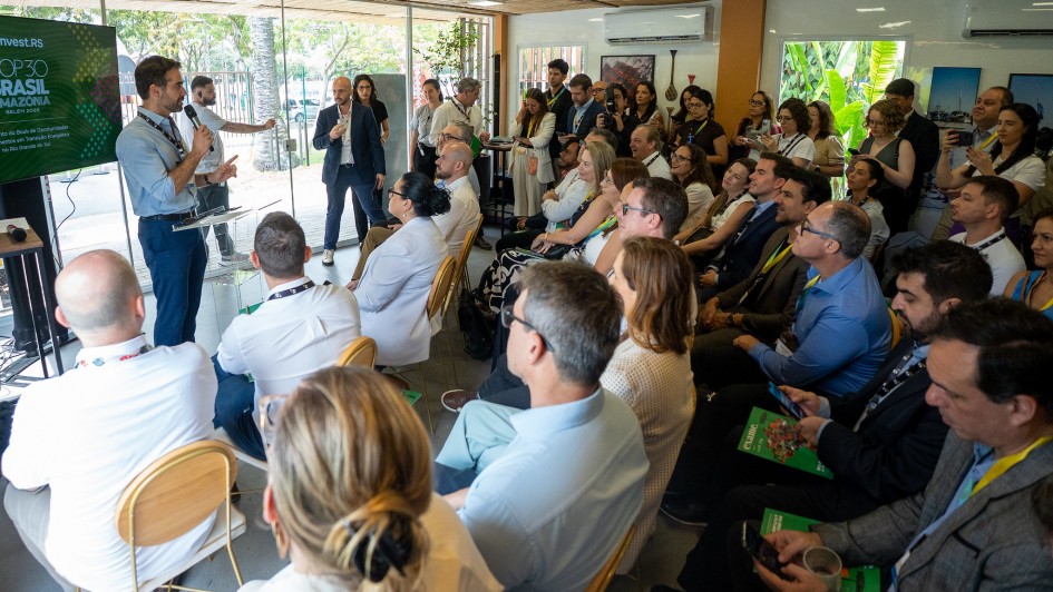 Ambiente interno com plateia atenta a uma apresentação. O governador Eduardo Leite aparece à esquerda, falando ao microfone e gesticulando enquanto apresenta os investimentos do Rio Grande do Sul durante a COP30. Ao fundo, um telão exibe o tema “Invest.RS — COP30 Brasil Amazônia”.