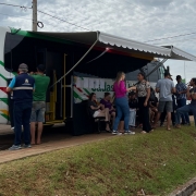 A imagem exibe um ônibus grande, branco e personalizado, estacionado em uma área gramada ao ar livre, servindo como unidade móvel de atendimento.

Ônibus/Unidade Móvel: O veículo está de lado, com a traseira levemente visível à esquerda. Ele é predominantemente branco com detalhes em listras verdes, vermelhas e amarelas. O logo do Governo do Estado do Rio Grande do Sul e a inscrição "Cadastro Único" estão visíveis na lateral. O lado direito do ônibus está totalmente aberto, com uma marquise branca e verde estendida, criando uma área coberta para atendimento.

Pessoas e Atendimento: Um grupo de cerca de dez pessoas (homens e mulheres) está reunido sob a marquise ou em pé próximo à entrada aberta do ônibus. Algumas pessoas estão sentadas em cadeiras dobráveis, enquanto outras conversam em pé. Um homem de coshirt azul e colete de segurança azul (possivelmente um atendente) está de costas para a câmera, próximo à lateral do ônibus.

Ambiente: O ônibus está estacionado em uma área elevada, com um talude gramado em primeiro plano. O fundo mostra uma paisagem de água (possivelmente um rio, lago ou mar, embora não seja claro) e uma palmeira à direita. O céu está nublado, com nuvens cinzentas e brancas. No canto inferior direito, parte de um veículo estacionado está visível.