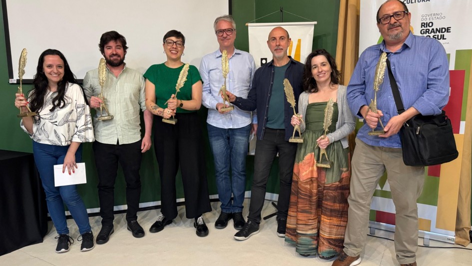 A imagem mostra sete pessoas em pé, posando para a foto e segurando o troféu do prêmio Prêmio Minuano de Literatura 2025. 