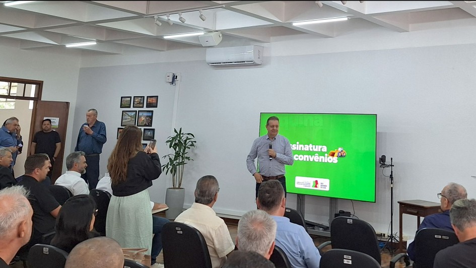 A imagem mostra um homem falando  ao microfone, em frente a um grupo de pessoas sentadas. Ao fundo, há um telão onde se lê "Assinatura de convênios" e as marcas da Secretaria da Agricultura e do Governo do RS. 