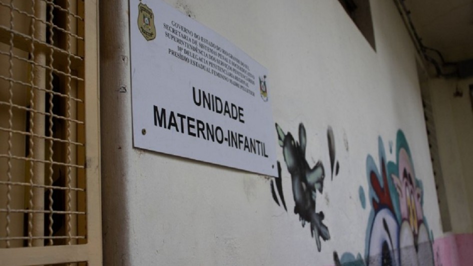 Uma foto em ambiente interno que captura uma placa de sinalização de uma unidade prisional. A placa, de cor branca com texto em letras pretas, está fixada em uma parede de cor clara (bege ou branca suja). O texto central é "UNIDADE MATERNO-INFANTIL".

No topo da placa, há o logotipo do governo do Rio Grande do Sul e um texto menor que identifica a instituição: "GOVERNO DO ESTADO DO RIO GRANDE DO SUL" seguido por "SECRETARIA DE SISTEMA PENAL E SOCIOEDUCATIVO", "SUPERINTENDÊNCIA DOS SERVIÇOS PENITENCIÁRIOS", e o nome da unidade: "PRISÃO ESTADUAL FEMININA MADRE PELLETIER" (o final é cortado).

À direita da placa, há uma pintura de rua ou grafite com figuras escuras e abstratas, com detalhes em cores claras, rosa e azul/turquesa. No canto esquerdo, parte de uma grade ou portão de metal do sistema prisional é visível, indicando o ambiente. A foto foi tirada em ângulo para dar destaque à placa.