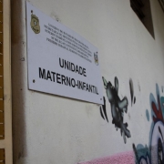 Uma foto em ambiente interno que captura uma placa de sinalização de uma unidade prisional. A placa, de cor branca com texto em letras pretas, está fixada em uma parede de cor clara (bege ou branca suja). O texto central é "UNIDADE MATERNO-INFANTIL".

No topo da placa, há o logotipo do governo do Rio Grande do Sul e um texto menor que identifica a instituição: "GOVERNO DO ESTADO DO RIO GRANDE DO SUL" seguido por "SECRETARIA DE SISTEMA PENAL E SOCIOEDUCATIVO", "SUPERINTENDÊNCIA DOS SERVIÇOS PENITENCIÁRIOS", e o nome da unidade: "PRISÃO ESTADUAL FEMININA MADRE PELLETIER" (o final é cortado).

À direita da placa, há uma pintura de rua ou grafite com figuras escuras e abstratas, com detalhes em cores claras, rosa e azul/turquesa. No canto esquerdo, parte de uma grade ou portão de metal do sistema prisional é visível, indicando o ambiente. A foto foi tirada em ângulo para dar destaque à placa.