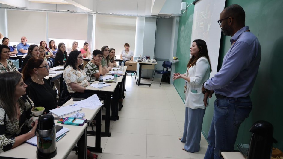 Foto em sala de aula de um homem e uma mulher falando em frente à turma, composta por professores sentados em classes.
