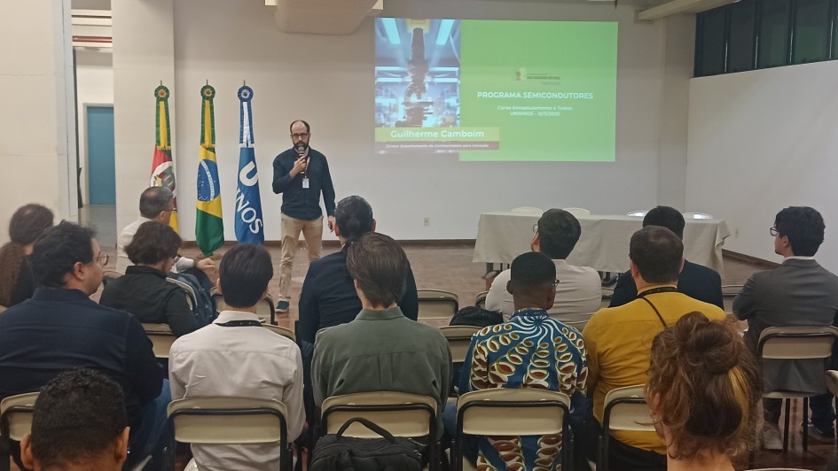 Uma foto em ambiente interno, em uma sala de aula ou audit&oacute;rio. Um homem (Guilherme Cambeim), no centro, est&aacute; de p&eacute; em um estrado de madeira, falando em um microfone que segura na m&atilde;o direita, voltado para a plateia. Ele veste uma camisa polo escura, cal&ccedil;a c&aacute;qui e &oacute;culos.

Atr&aacute;s dele, uma tela de proje&ccedil;&atilde;o exibe um slide com o t&iacute;tulo "PROGRAMA SEMICONDUTORES", um subt&iacute;tulo sobre cursos e uma foto vertical com foco em um equipamento ou placa de circuito. O nome "Guilherme Cambeim" aparece em letras menores abaixo.

A plateia &eacute; composta por cerca de 20 pessoas (a maioria de costas para a c&acirc;mera), sentadas em cadeiras brancas em fileiras. No lado esquerdo do estrado, h&aacute; tr&ecirc;s mastros com bandeiras: a do Rio Grande do Sul, a do Brasil e uma bandeira com o logotipo da ONU/UNOS. A sala tem paredes brancas e tetos baixos com ilumina&ccedil;&atilde;o de LED embutida.