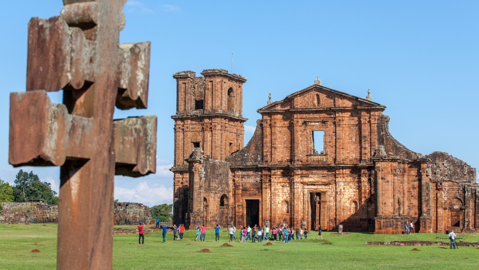 A imagem mostra as ruínas de uma antiga construção missioneira em pedra avermelhada, situadas em um amplo gramado verde. No primeiro plano, um grande crucifixo de madeira aparece desfocado, enquanto ao fundo um grupo de visitantes caminha pelo sítio histórico. O céu está limpo e azul, compondo uma cena típica de turismo cultural ao ar livre.