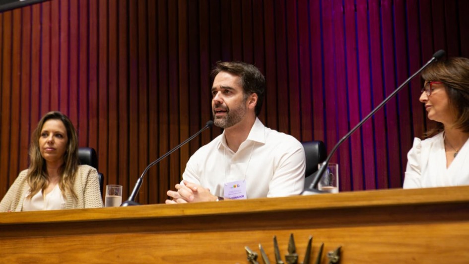 Uma foto em ambiente interno, em um fórum ou encontro. A foto foca em um painel de discussão no palco. Três pessoas estão sentadas atrás de uma bancada de madeira escura com um brasão ou emblema. No centro, o homem (Eduardo Leite), vestindo uma camisa social branca, está inclinado para a frente, com as mãos entrelaçadas sobre o púlpito, olhando para a frente enquanto fala em um microfone.

À sua esquerda, uma mulher de casaco bege e blusa branca está sentada, observando. À sua direita, outra mulher de óculos, blusa branca e blazer escuro escuta atenta, com um microfone à sua frente. As luzes de fundo projetam tons de roxo e rosa em painéis verticais de madeira escura, criando um contraste e um ambiente de evento formal.