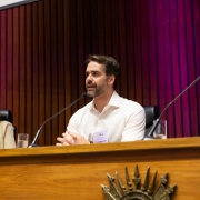 Uma foto em ambiente interno, em um fórum ou encontro. A foto foca em um painel de discussão no palco. Três pessoas estão sentadas atrás de uma bancada de madeira escura com um brasão ou emblema. No centro, o homem (Eduardo Leite), vestindo uma camisa social branca, está inclinado para a frente, com as mãos entrelaçadas sobre o púlpito, olhando para a frente enquanto fala em um microfone.

À sua esquerda, uma mulher de casaco bege e blusa branca está sentada, observando. À sua direita, outra mulher de óculos, blusa branca e blazer escuro escuta atenta, com um microfone à sua frente. As luzes de fundo projetam tons de roxo e rosa em painéis verticais de madeira escura, criando um contraste e um ambiente de evento formal.