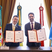 Uma foto de dois homens, o governador do Rio Grande do Sul (à direita, de terno escuro) e o governador da província japonesa de Shiga (à esquerda, de terno e gravata dourada), em pé, posando juntos com cópias abertas de uma declaração conjunta. Ambos sorriem para a câmera e seguram os documentos com as mãos nas laterais e no centro.

A cópia do documento que cada um segura é idêntica, apresentando um texto em inglês no lado esquerdo e em japonês (ou português, dependendo do lado) no lado direito, emoldurado por uma borda vermelha.

Atrás deles, há uma janela central com cortinas grossas de veludo marrom emoldurando-a. Nos cantos, estão hastes de bandeiras ornamentais com fitas coloridas (vermelho, verde e azul) e emblemas redondos: à esquerda, as cores e o brasão do Rio Grande do Sul e, à direita, uma bandeira de cor azul claro e branco.

A mesa em primeiro plano possui placas de identificação com os nomes dos líderes: "TAZO MIKAZUKI" e "EDUARDO LEITE". A cena é formal, em ambiente de reunião de alto nível governamental.