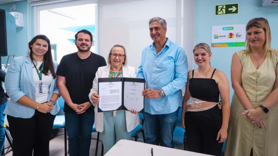 Uma foto em close-up de uma cerimônia de assinatura de convênio em Campo Bom. Seis pessoas (quatro mulheres e dois homens) estão alinhadas, sorrindo e posando atrás de uma mesa branca.

No centro, uma mulher mais velha, de cabelo grisalho e blazer branco sobre calças azul-claro, e um homem alto, de cabelos grisalhos e camisa social azul-clara (manga comprida) e jeans, estão segurando uma pasta preta aberta contendo o documento do convênio. O homem e a mulher segurando a pasta parecem ser as autoridades principais no centro do enquadramento.

À esquerda, duas mulheres vestem blazers azul-claro e escuro, sendo que a primeira usa um crachá pendurado. À direita, uma mulher de blusa preta e saia/calça escura e, ao lado dela, outra mulher de vestido ou casaco longo bege completam o grupo.

A sala tem paredes de cor clara (azul-bebê e branco). No fundo, há uma janela grande e esquadrias azuis que dão para uma área externa. Na parede do fundo, há sinais de segurança ("Saída de Emergência" com seta verde) e um cartaz colorido de aviso ou informação, com o logotipo do governo do Rio Grande do Sul.