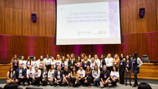 Um grande grupo de participantes posa para uma foto coletiva no auditório, em frente ao palco onde é projetado o título do evento “1º Encontro Estadual Mulheres para o Mundo – Redes de Proteção da Mulher”. As pessoas estão alinhadas em várias fileiras, sorrindo para a câmera.