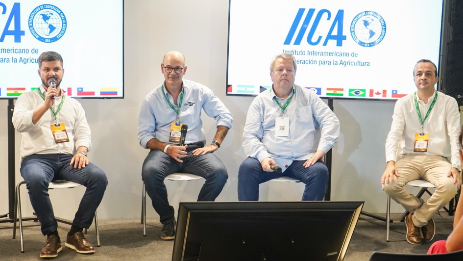 Uma foto em plano médio de quatro homens sentados em cadeiras brancas em um palco de conferência, participando de um painel de discussão focado em tecnologia agrícola.

Da esquerda para a direita:

O primeiro homem (de camisa branca, blazer escuro, jeans e botas marrons) segura um microfone e gesticula enquanto fala.

O segundo homem (de camisa social azul-clara, óculos e jeans) está atento, com as mãos sobre as pernas.

O terceiro homem (de camisa branca e jeans) está sentado de forma relaxada, segurando um microfone.

O quarto homem (de camisa social branca e calças cáqui) está sentado com as pernas cruzadas.

Atrás dos palestrantes, duas grandes telas verticais (painéis de LED ou projeções) exibem o logotipo do IICA ("Instituto Interamericano de Cooperação para a Agricultura") e bandeiras de vários países. Um monitor preto horizontal está no chão, em frente aos palestrantes. A direita, a lateral de uma mulher em primeiro plano, sentada na audiência, é visível.