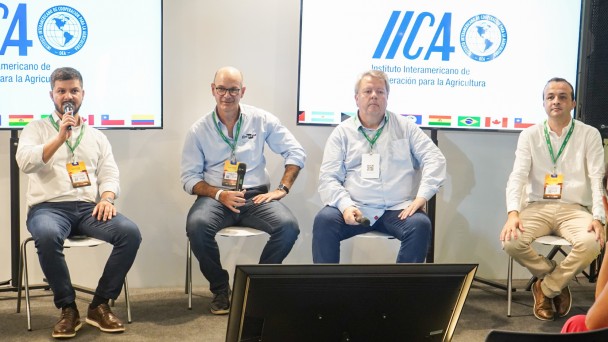 Uma foto em plano médio de quatro homens sentados em cadeiras brancas em um palco de conferência, participando de um painel de discussão focado em tecnologia agrícola.

Da esquerda para a direita:

O primeiro homem (de camisa branca, blazer escuro, jeans e botas marrons) segura um microfone e gesticula enquanto fala.

O segundo homem (de camisa social azul-clara, óculos e jeans) está atento, com as mãos sobre as pernas.

O terceiro homem (de camisa branca e jeans) está sentado de forma relaxada, segurando um microfone.

O quarto homem (de camisa social branca e calças cáqui) está sentado com as pernas cruzadas.

Atrás dos palestrantes, duas grandes telas verticais (painéis de LED ou projeções) exibem o logotipo do IICA ("Instituto Interamericano de Cooperação para a Agricultura") e bandeiras de vários países. Um monitor preto horizontal está no chão, em frente aos palestrantes. A direita, a lateral de uma mulher em primeiro plano, sentada na audiência, é visível.