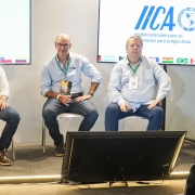Uma foto em plano médio de quatro homens sentados em cadeiras brancas em um palco de conferência, participando de um painel de discussão focado em tecnologia agrícola.

Da esquerda para a direita:

O primeiro homem (de camisa branca, blazer escuro, jeans e botas marrons) segura um microfone e gesticula enquanto fala.

O segundo homem (de camisa social azul-clara, óculos e jeans) está atento, com as mãos sobre as pernas.

O terceiro homem (de camisa branca e jeans) está sentado de forma relaxada, segurando um microfone.

O quarto homem (de camisa social branca e calças cáqui) está sentado com as pernas cruzadas.

Atrás dos palestrantes, duas grandes telas verticais (painéis de LED ou projeções) exibem o logotipo do IICA ("Instituto Interamericano de Cooperação para a Agricultura") e bandeiras de vários países. Um monitor preto horizontal está no chão, em frente aos palestrantes. A direita, a lateral de uma mulher em primeiro plano, sentada na audiência, é visível.