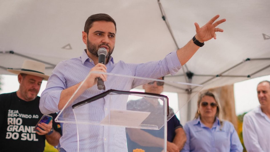Homem fala ao microfone em evento oficial, atrás de púlpito transparente com brasão. Pessoas acompanham sob tenda, e há mensagem de boas-vindas ao Rio Grande do Sul. Cena pública e institucional.