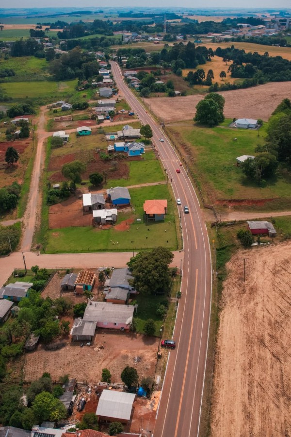 Vista aérea de área rural com estrada asfaltada, casas espaçadas e campos cultivados. Paisagem verde e tranquila.