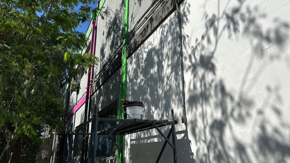 Uma foto em close-up de uma parede branca de um prédio em obras ou revitalização (possivelmente a TVE). A parede lisa está parcialmente coberta pelas sombras projetadas de galhos e folhas de árvores frondosas que estão do lado esquerdo da imagem, em primeiro plano.

A parede branca é marcada por linhas verticais em cores vibrantes, típicas de revitalização, sendo uma linha rosa-choque e outra verde-limão.

Em um plano intermediário, um andaime simples de metal escuro está montado junto à parede, com uma prancha de metal (ou madeira) horizontal. Sobre esta prancha, há um balde de tinta ou massa claro, com uma vassoura ou escova saindo dele. A parte superior do andaime também tem tubos pretos e cabos que correm horizontalmente na parede. O céu azul intenso, visível no topo da imagem, sugere um dia de sol forte.