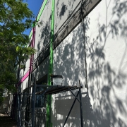 Uma foto em close-up de uma parede branca de um prédio em obras ou revitalização (possivelmente a TVE). A parede lisa está parcialmente coberta pelas sombras projetadas de galhos e folhas de árvores frondosas que estão do lado esquerdo da imagem, em primeiro plano.

A parede branca é marcada por linhas verticais em cores vibrantes, típicas de revitalização, sendo uma linha rosa-choque e outra verde-limão.

Em um plano intermediário, um andaime simples de metal escuro está montado junto à parede, com uma prancha de metal (ou madeira) horizontal. Sobre esta prancha, há um balde de tinta ou massa claro, com uma vassoura ou escova saindo dele. A parte superior do andaime também tem tubos pretos e cabos que correm horizontalmente na parede. O céu azul intenso, visível no topo da imagem, sugere um dia de sol forte.